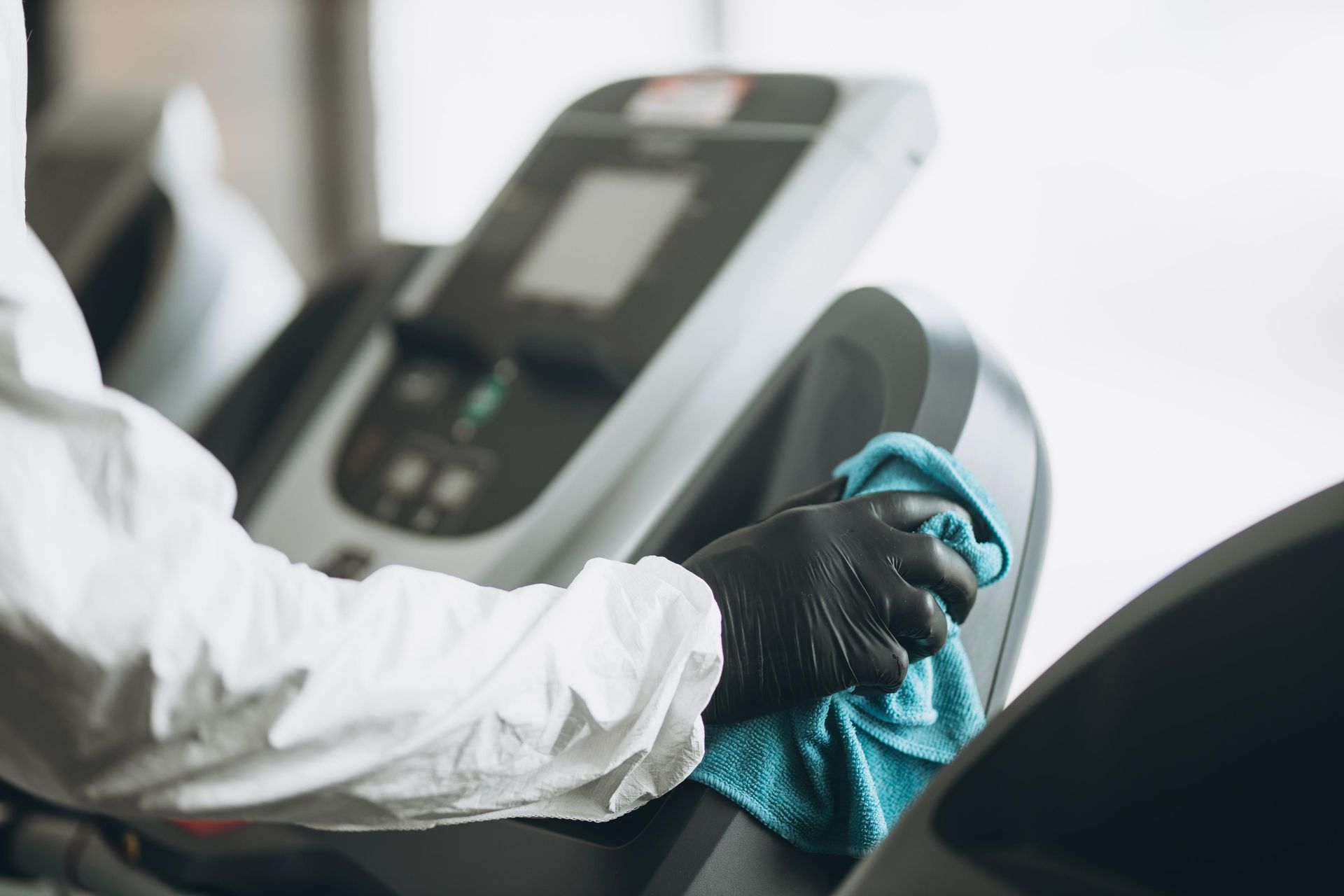 A person wearing a white protective suit and black gloves wipes down gym treadmill equipment with a blue cloth.
