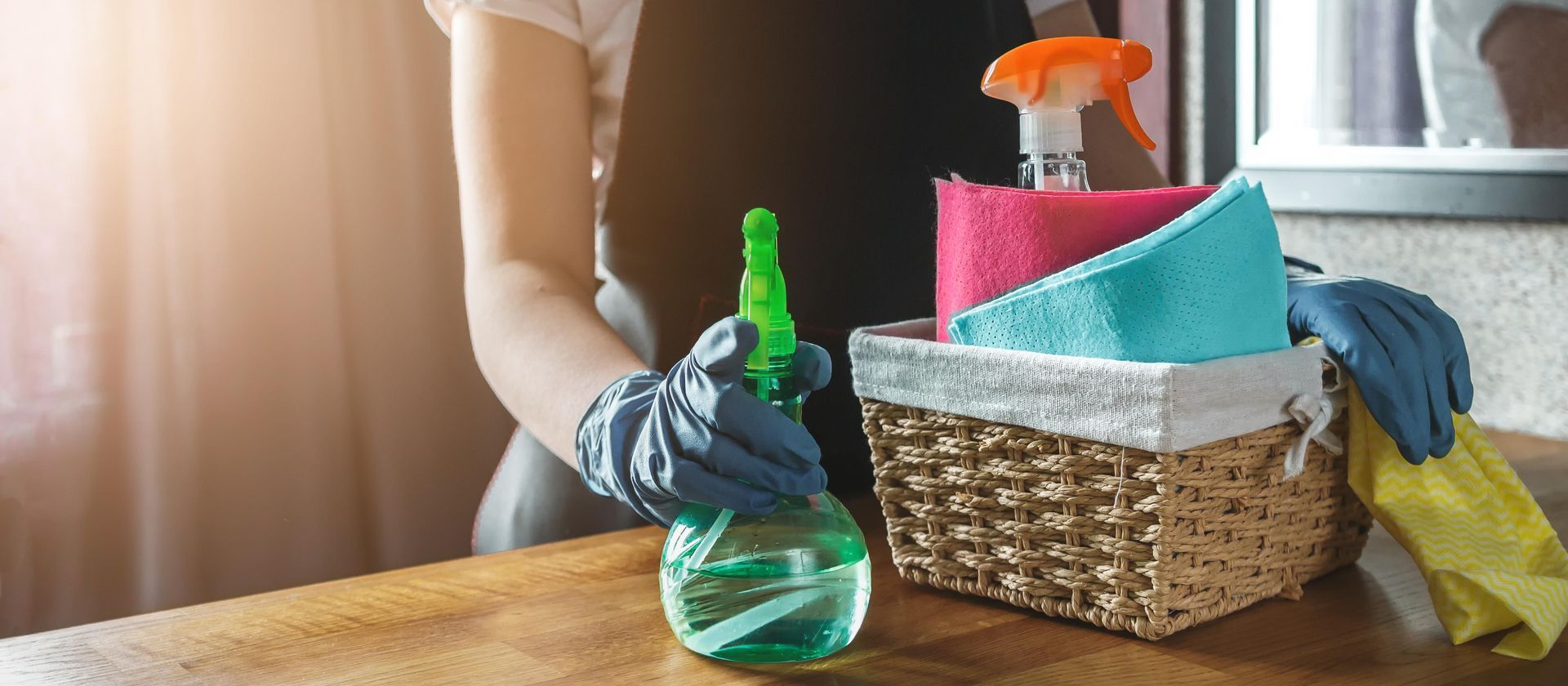 A person wearing blue gloves holds a spray bottle next to a wicker basket containing colorful cleaning cloths.