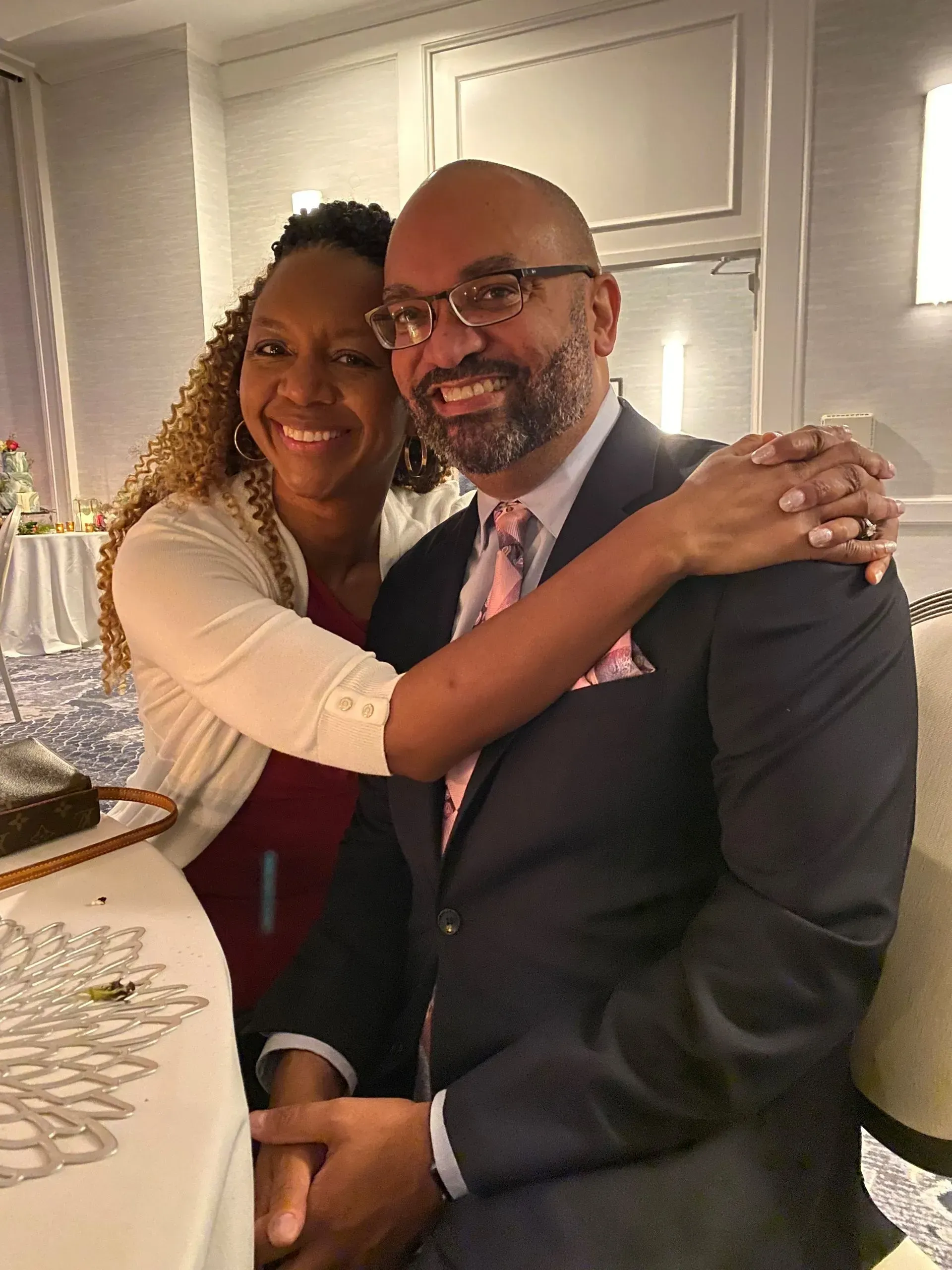 A smiling woman embraces a man wearing a suit and glasses, sitting at a table in a brightly lit, decorated room.