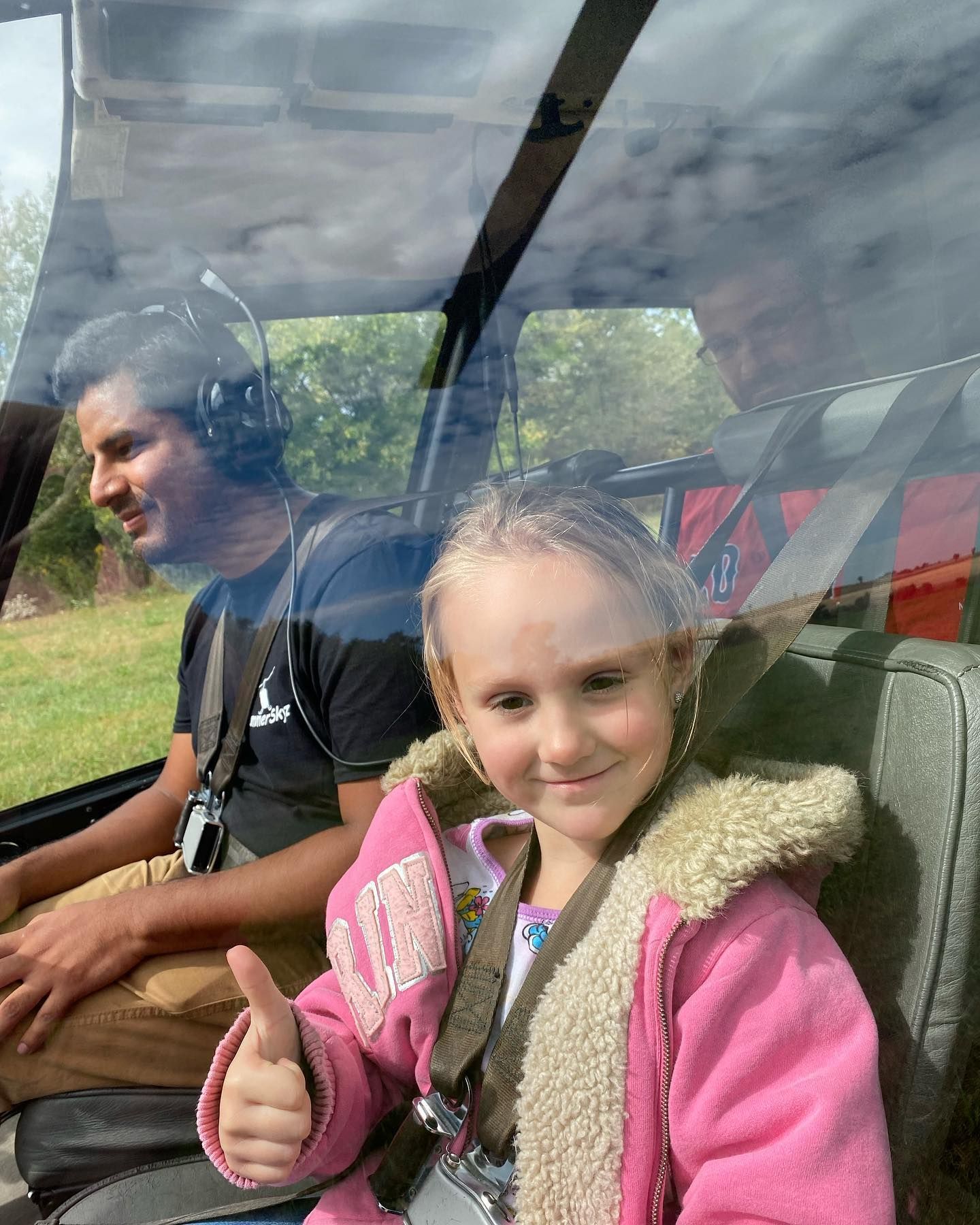 A smiling child in a pink jacket gives a thumbs-up in a helicopter, a man in a headset is at the controls.