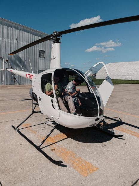 Helicopter with open door, two people inside. Outdoors on concrete. White and gray.