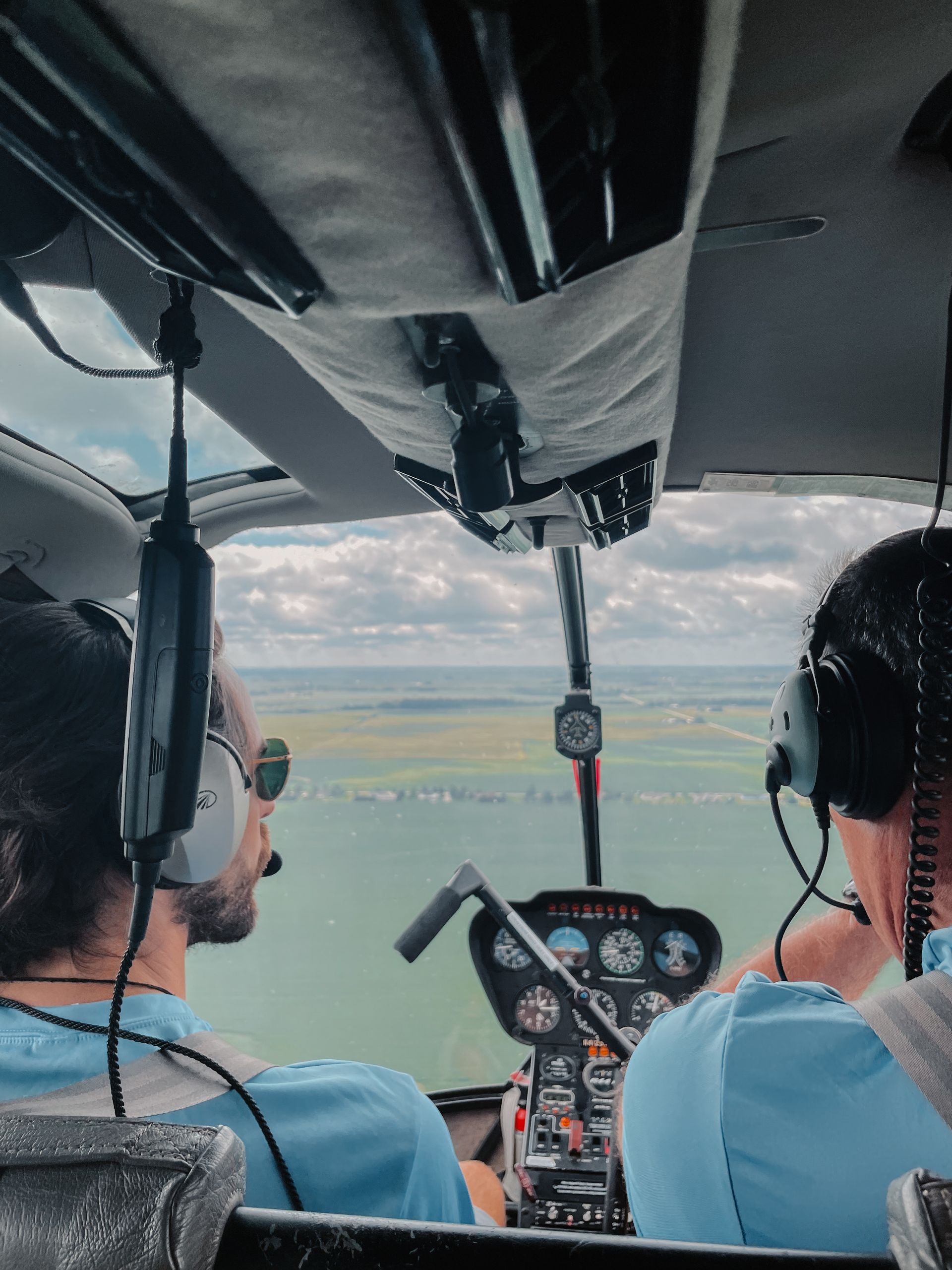 Two people in a helicopter cockpit flying over a green landscape with a cloudy sky.