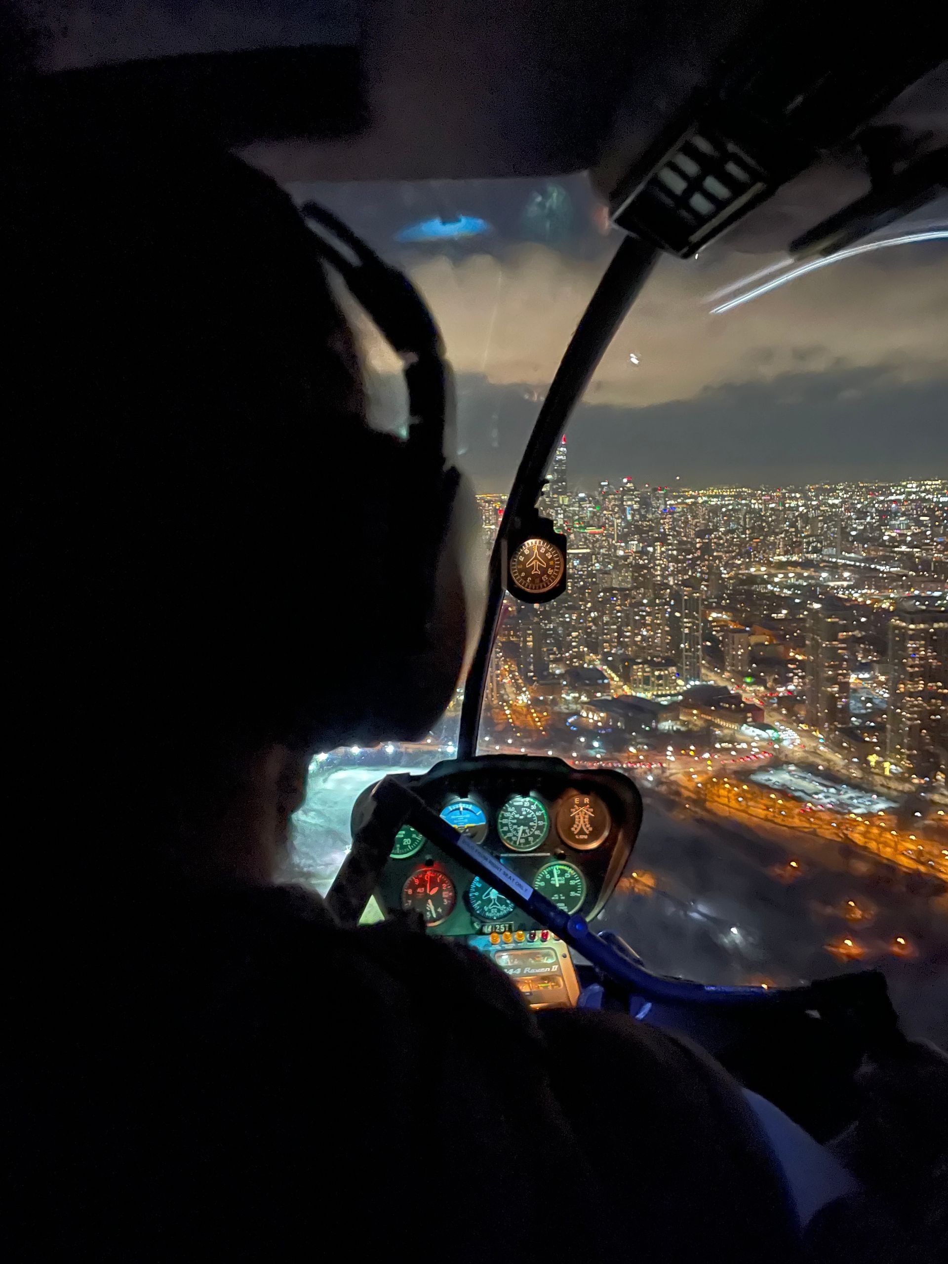 Pilot in helicopter cockpit at night, overlooking city lights.