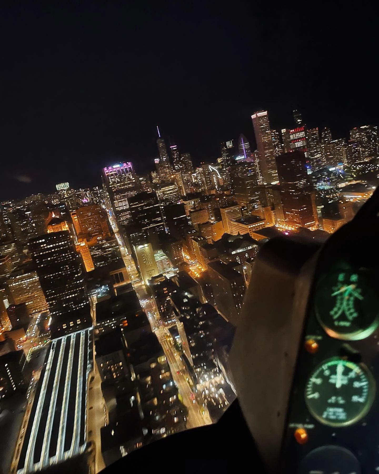 Nighttime aerial view of a brightly lit city skyline, seen from a cockpit with gauges visible.