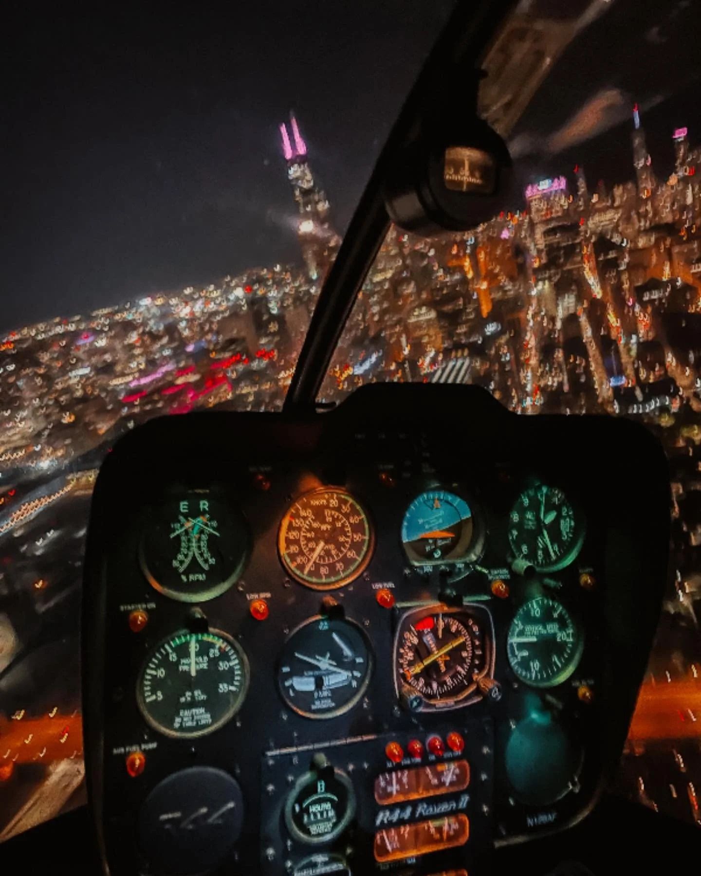 Helicopter cockpit view at night over a brightly lit city with skyscrapers. Instrument panel visible.