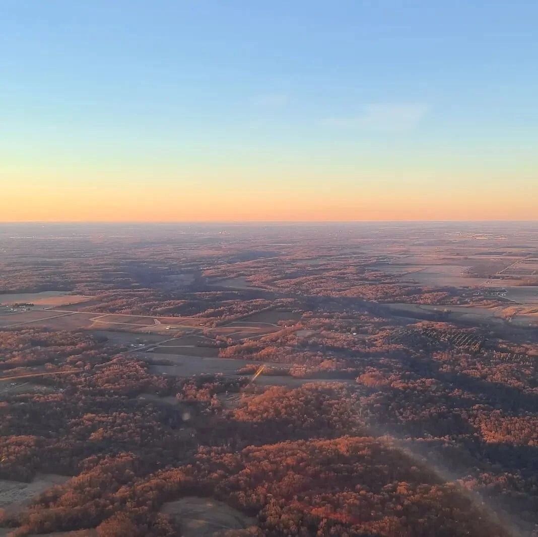 Aerial view of a hazy, forested landscape under a gradient sky of blue, orange, and yellow.