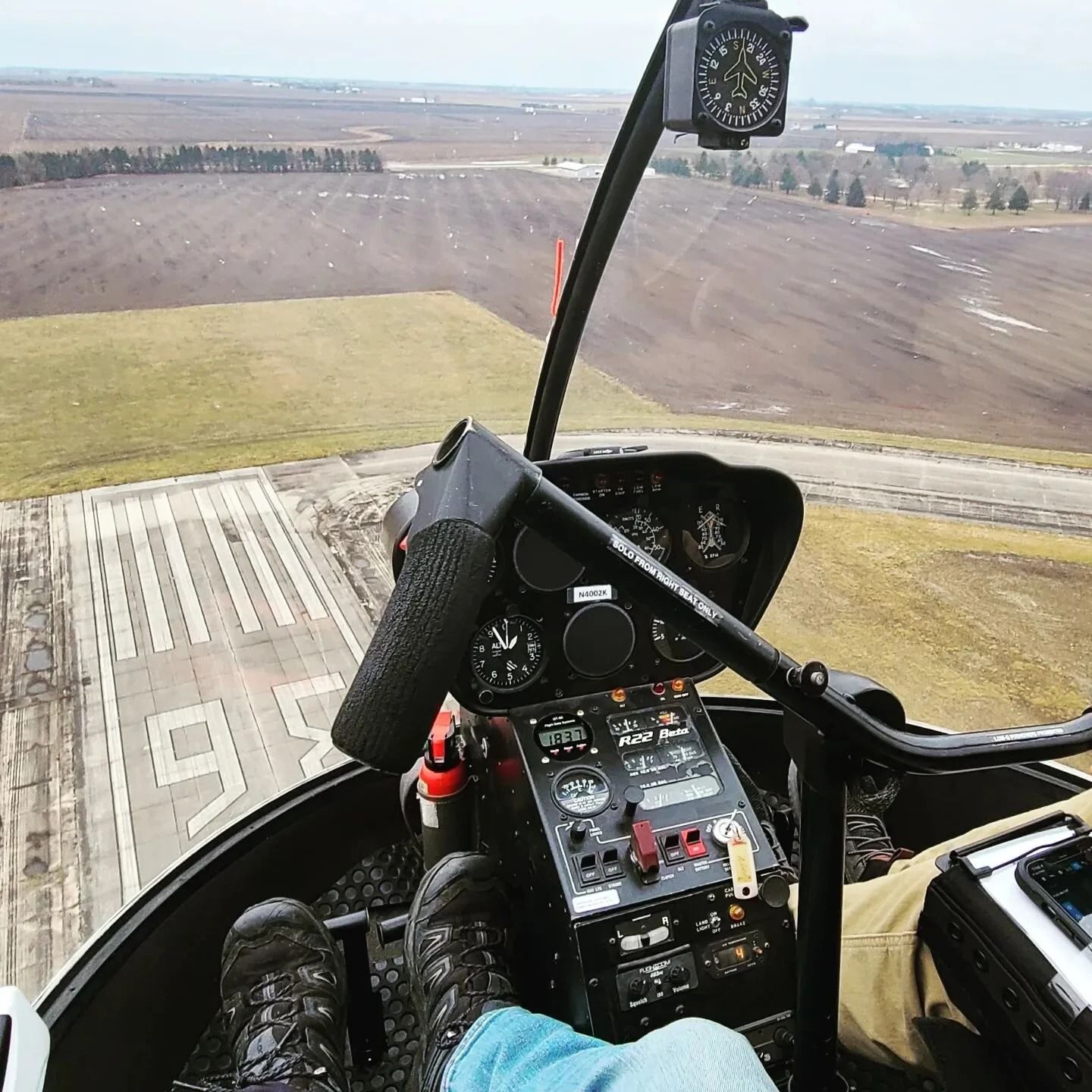 View from a helicopter cockpit looking down at a runway. Instrument panel visible with pilot's legs.