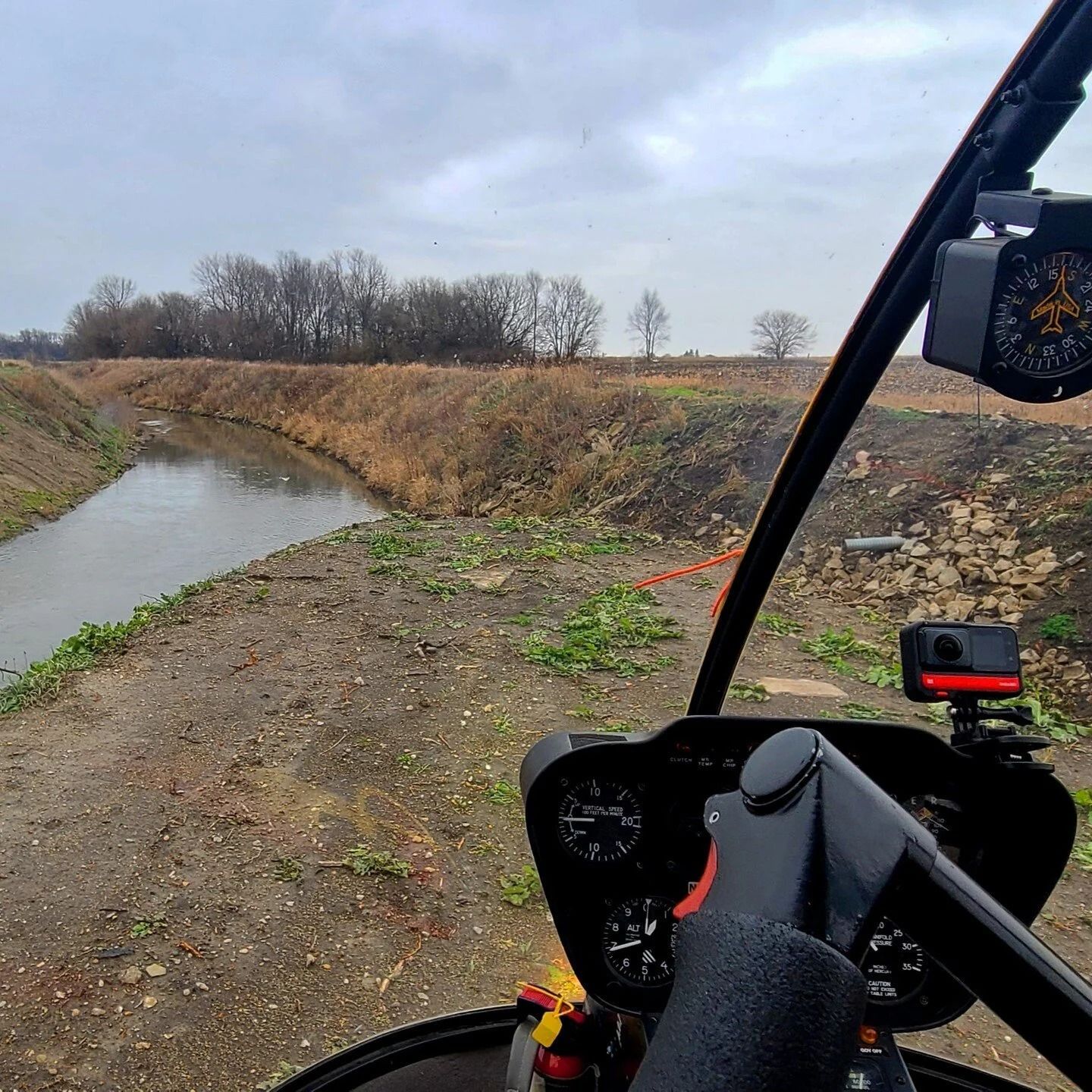 View from a helicopter cockpit looking at a waterway with trees on the horizon under a cloudy sky.