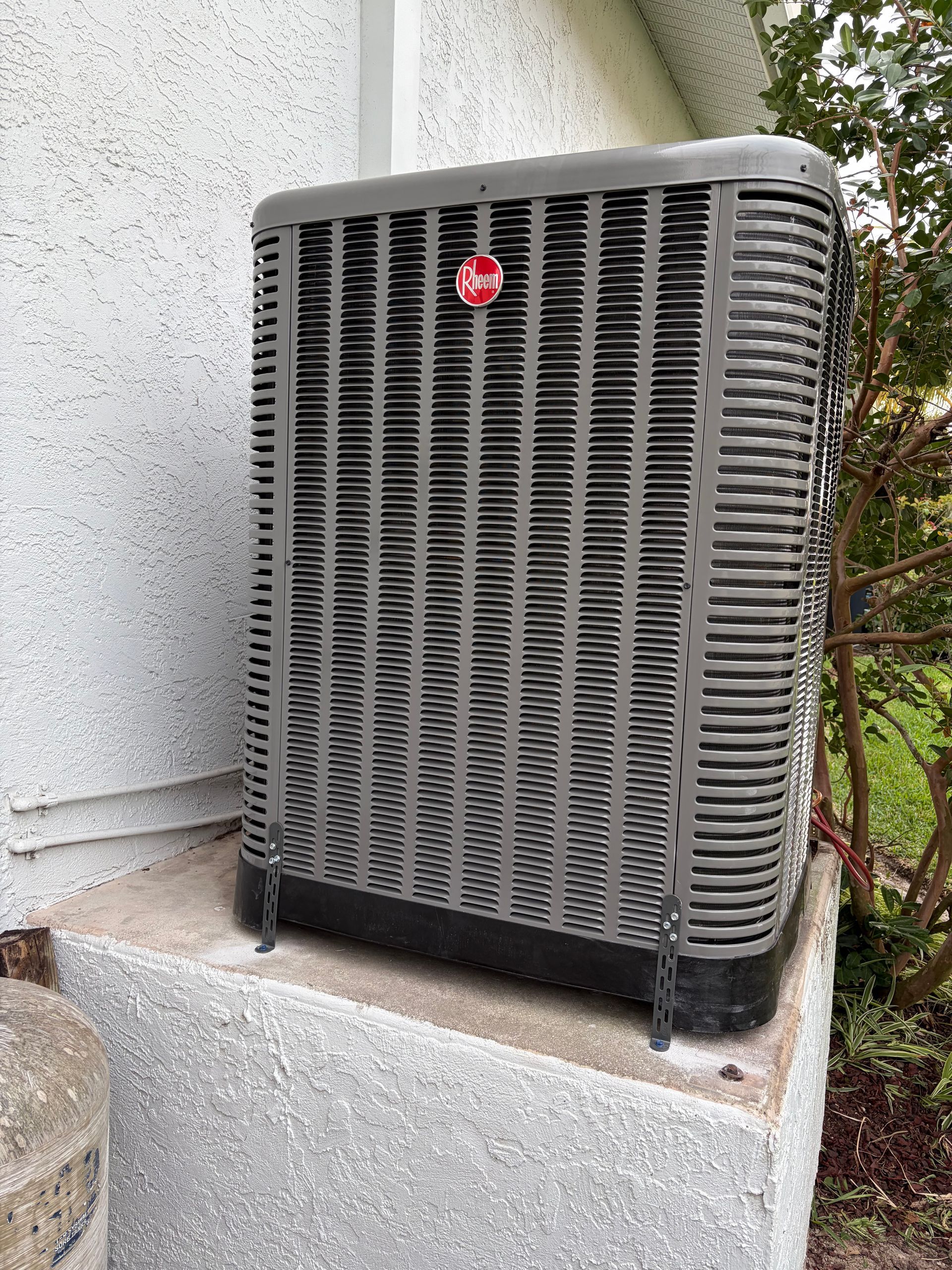 Outdoor air conditioning unit beside a house wall on a concrete pad, with a red logo on the front