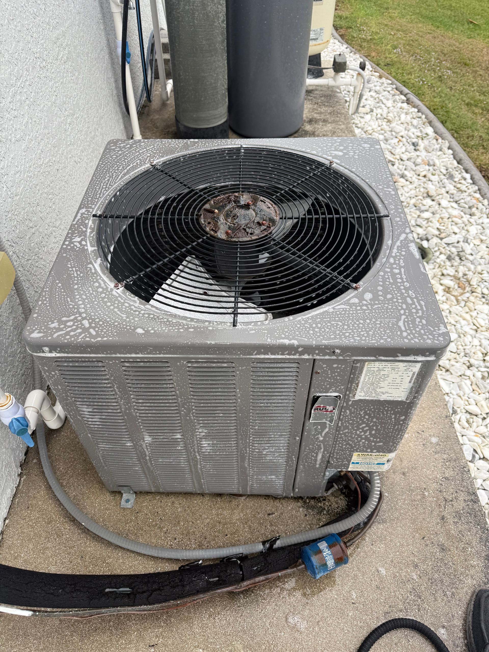 Outdoor HVAC condenser unit beside a building, with a top fan, hoses, and gravel ground around it