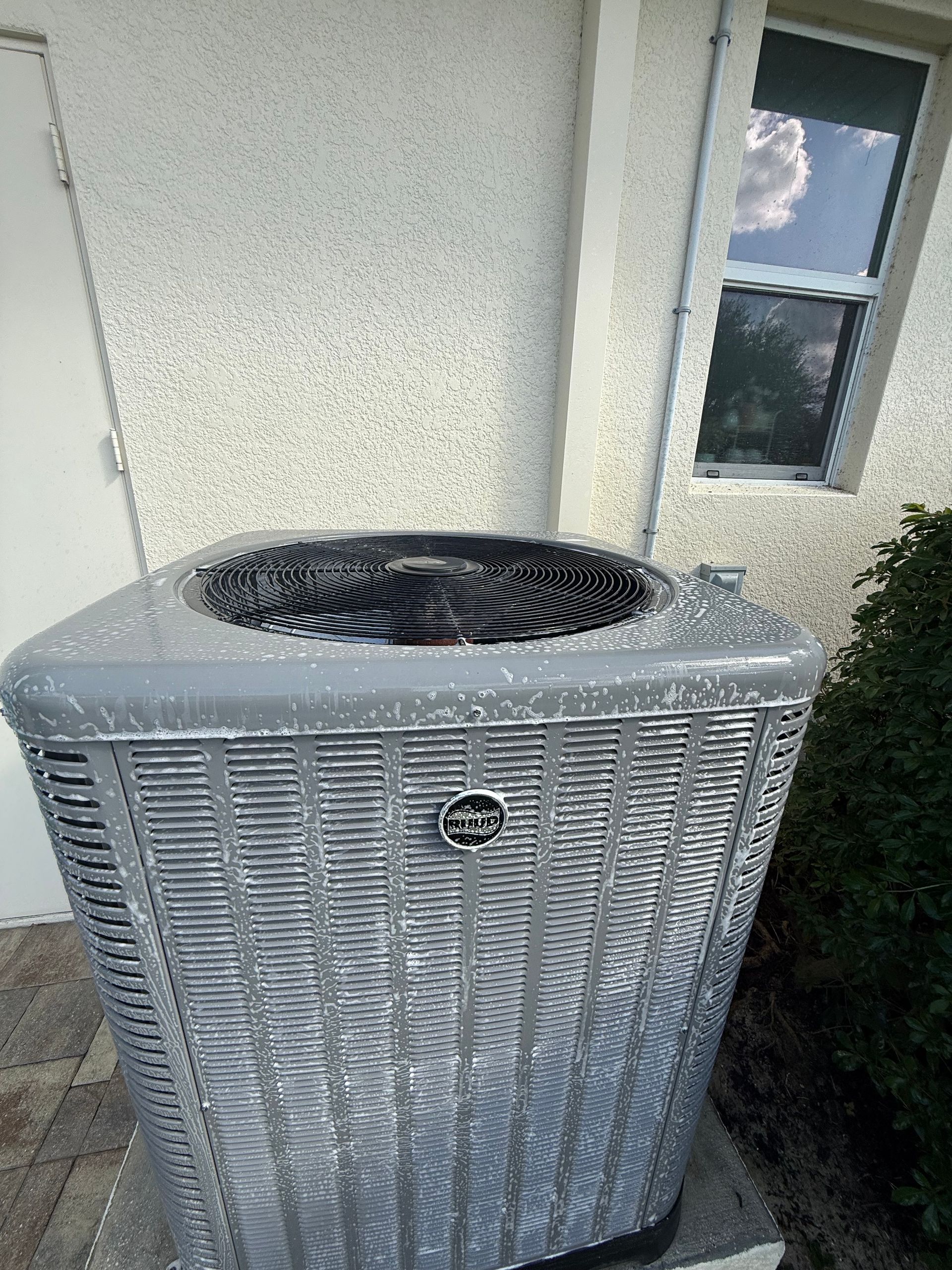 Outdoor air conditioning unit beside a house wall and window, with a fan on top.