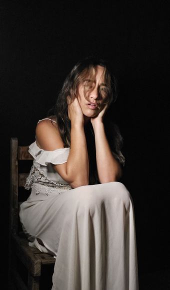 Uma mulher de vestido branco está sentada em uma cadeira com as mãos no rosto.