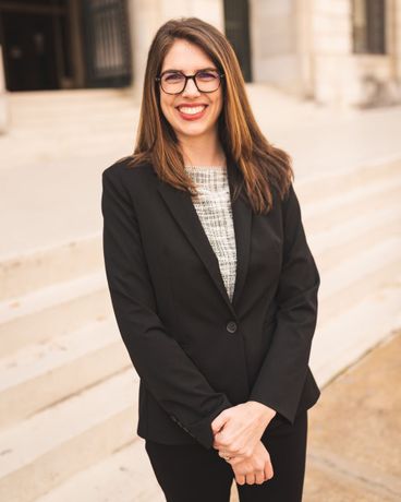 A woman wearing a black jacket and a pearl necklace is standing in front of a building.