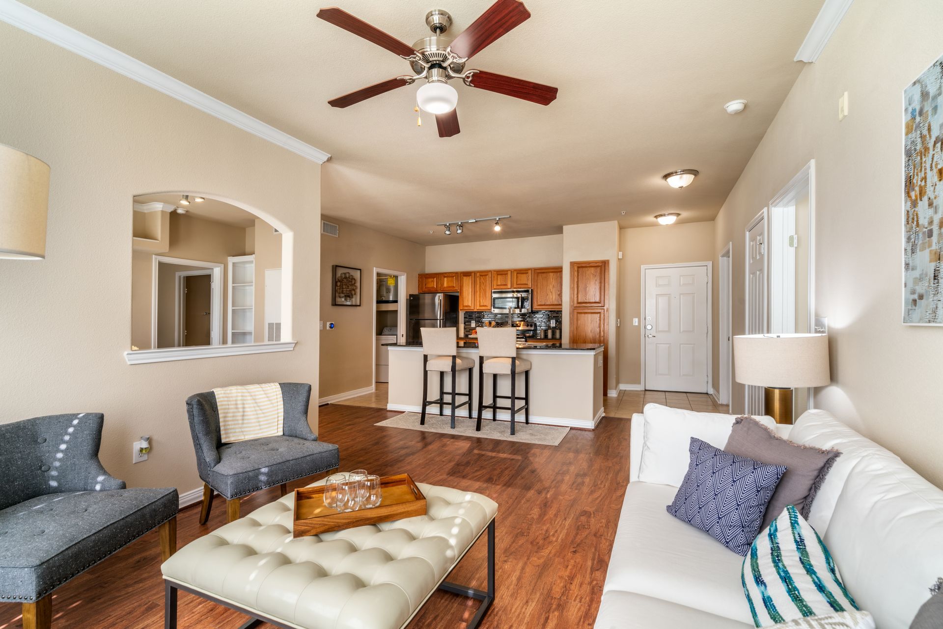Living room with kitchen in background; neutral colors, open layout, hardwood floor, and comfortable seating.