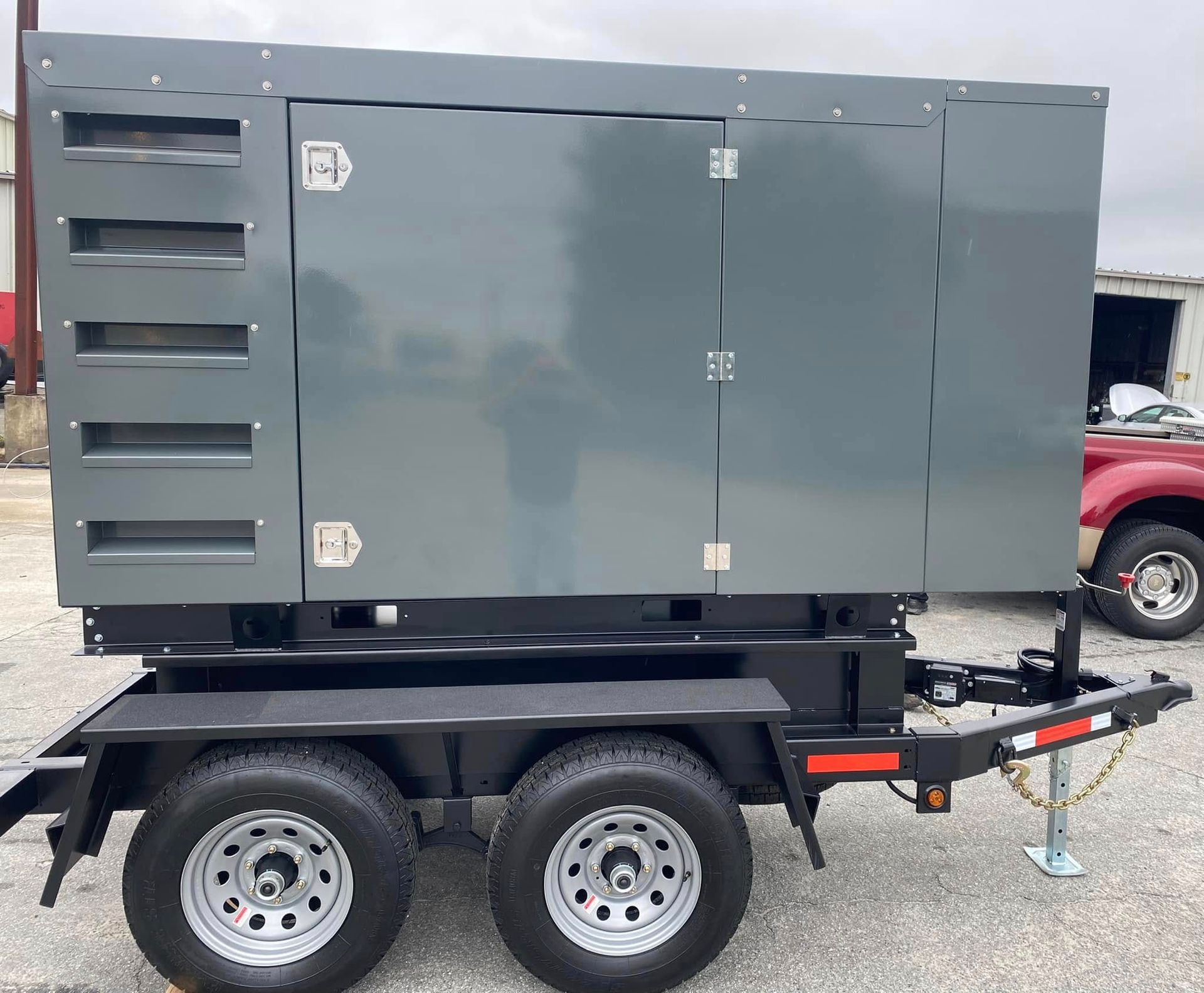 A large, gray industrial generator mounted on a black two-axle utility trailer parked on a gravel lot.