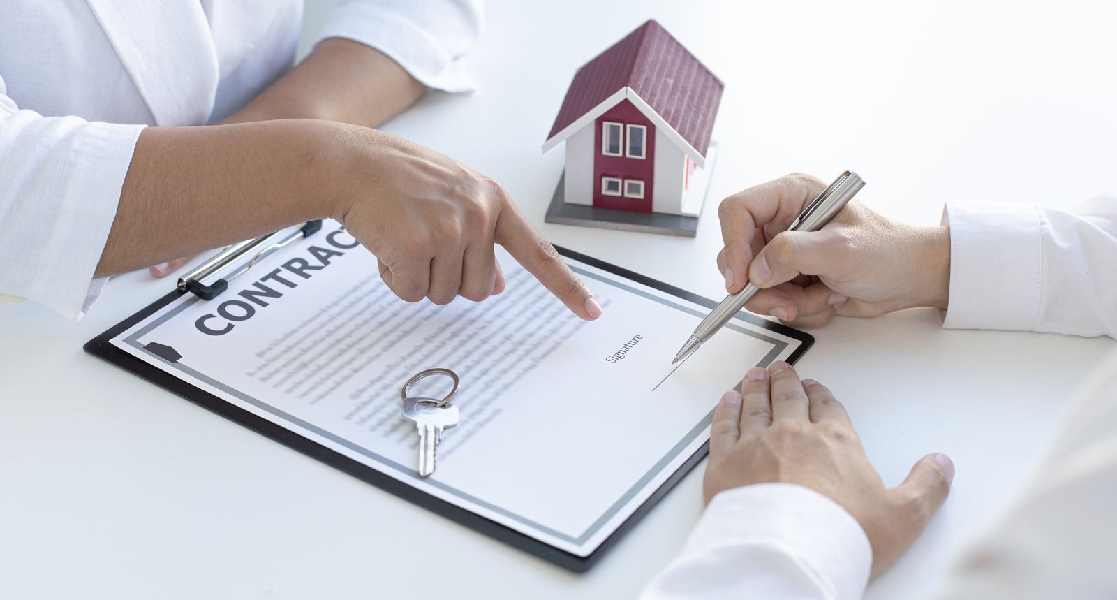 Two people sitting at a table with a contract, a small house model, and a key, one pointing to the signature line.