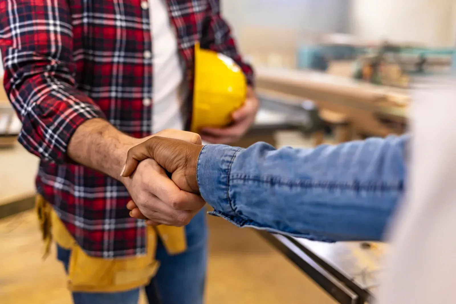 Two people in a woodworking shop shake hands, with one holding a yellow hard hat.
