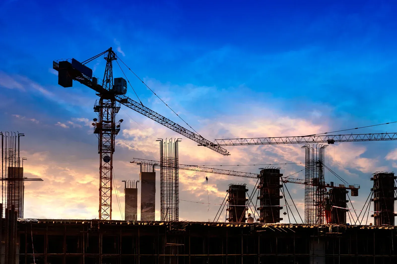 Silhouetted construction site at sunset, featuring cranes and unfinished vertical building structures against a blue sky.