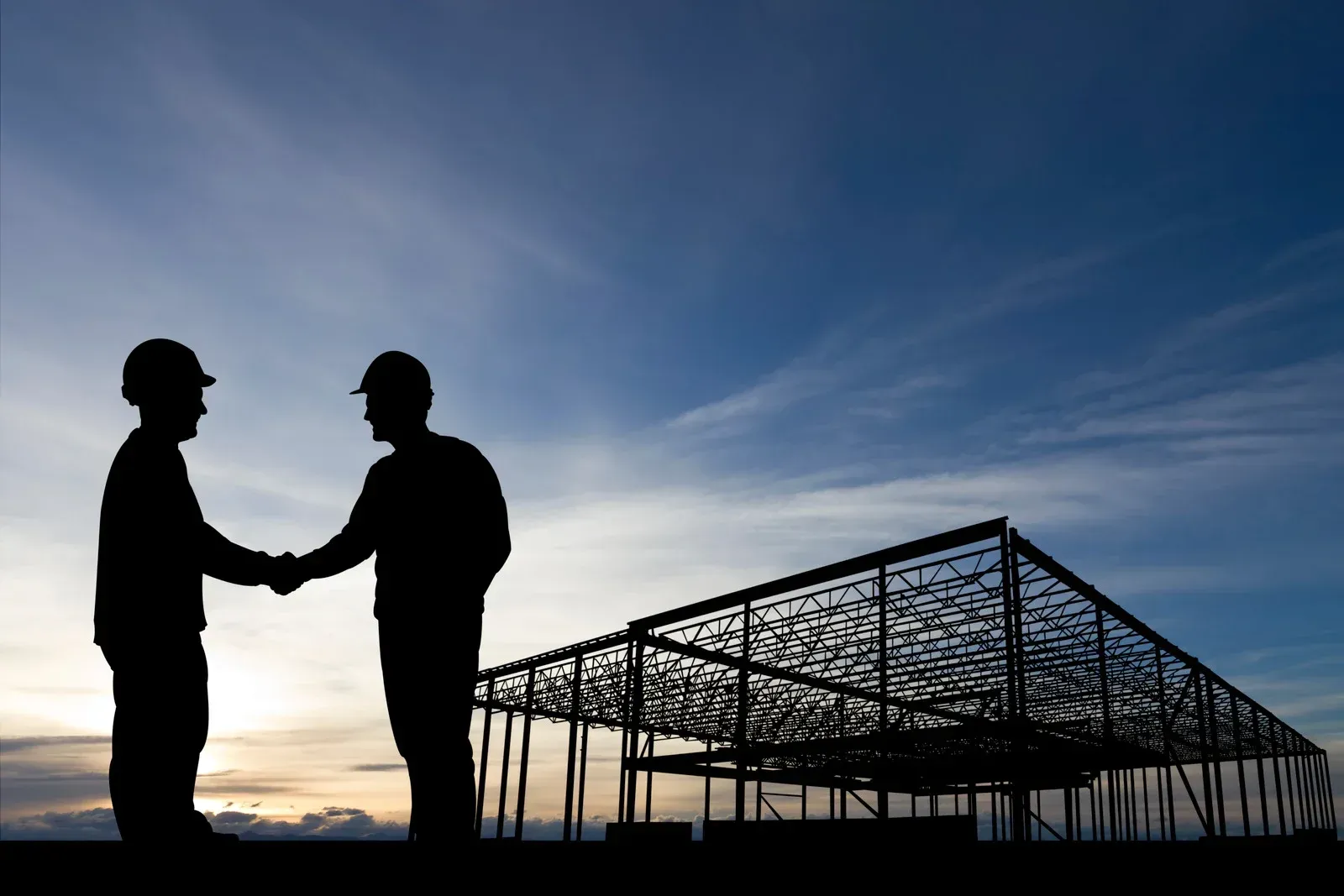Two workers in hard hats shake hands against a sunset sky beside a steel building framework.