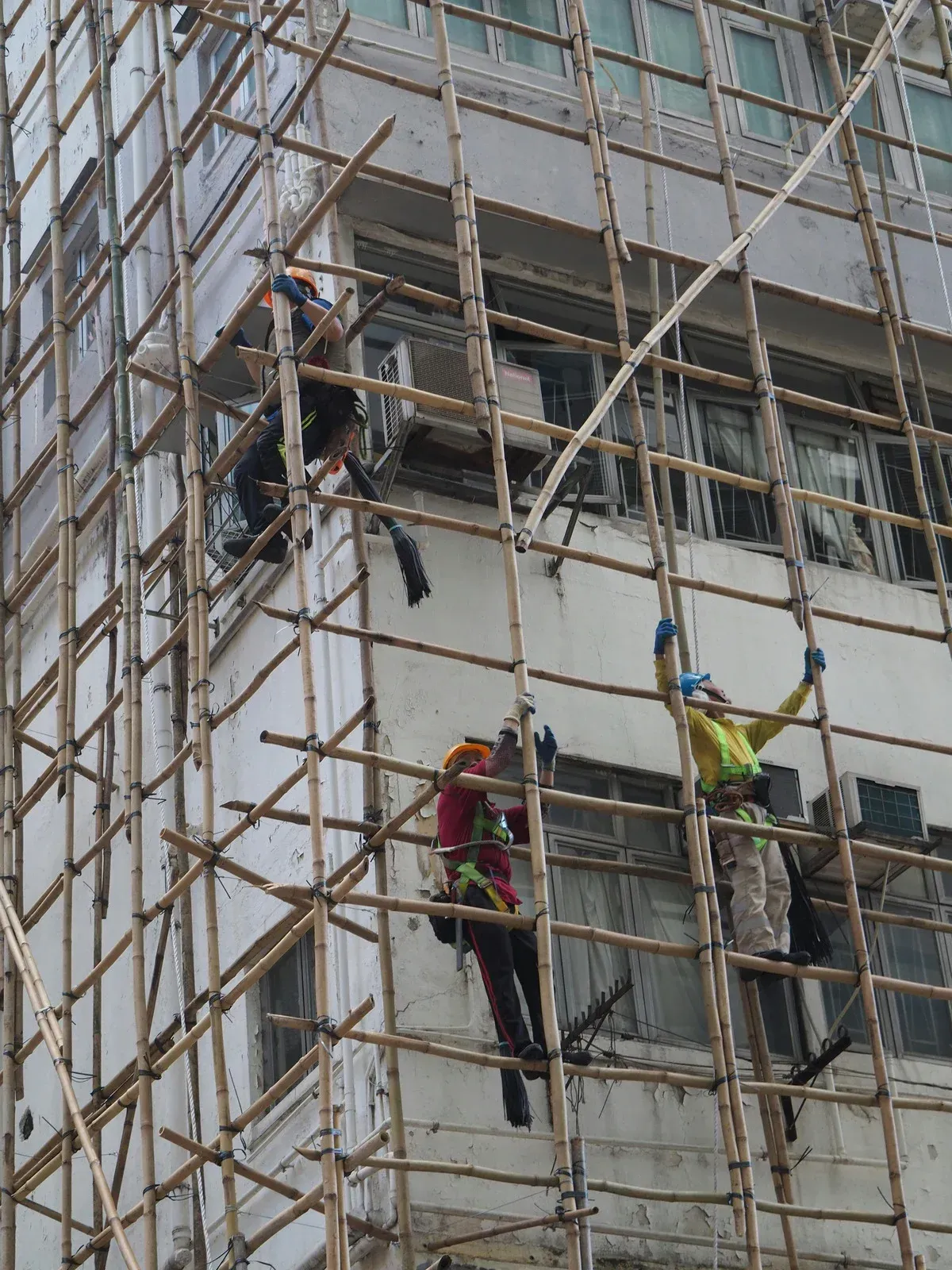 Three construction workers in safety gear climb a tall bamboo scaffolding system attached to a multi-story building.
