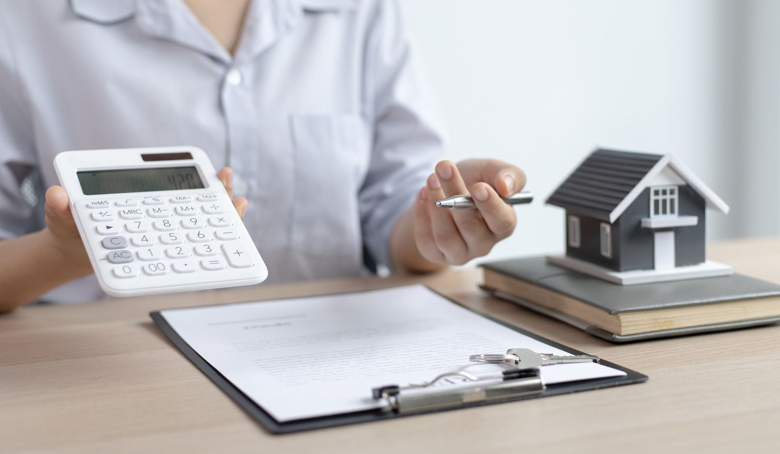 A person in a light shirt holds a calculator and pen over a clipboard with a model house on books nearby.