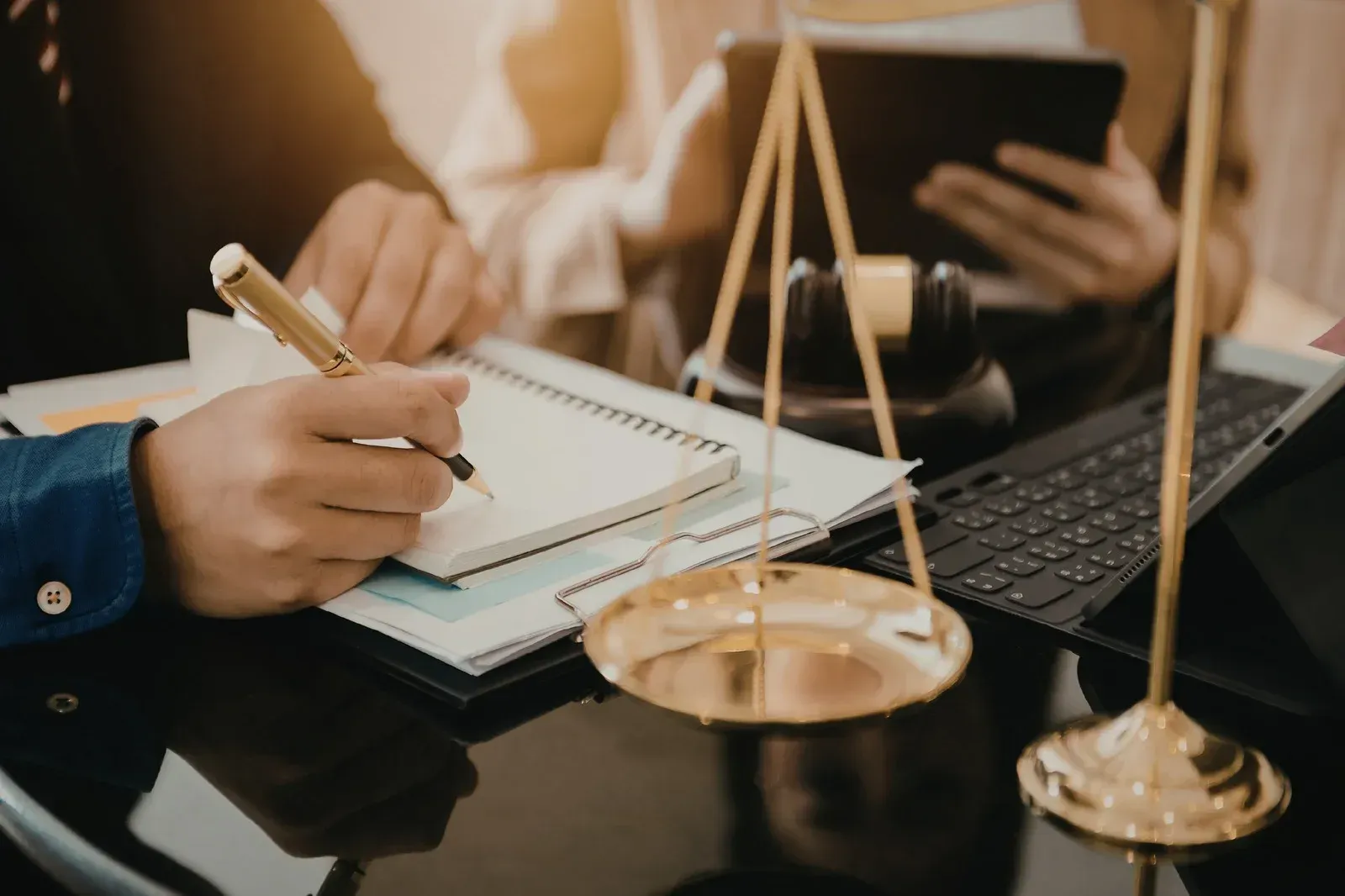 Close-up of a person writing in a notebook near a brass scales of justice, a gavel, and a tablet on a dark desk.