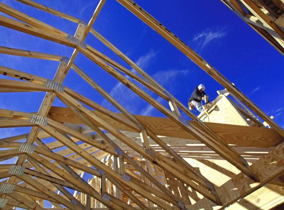 A construction worker installs roof trusses on a house frame under a bright blue sky.