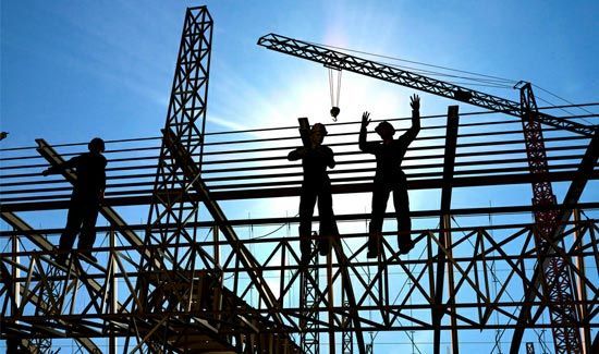 Silhouetted construction workers stand on a metal framework against a bright blue sky and sun, with cranes in the background.