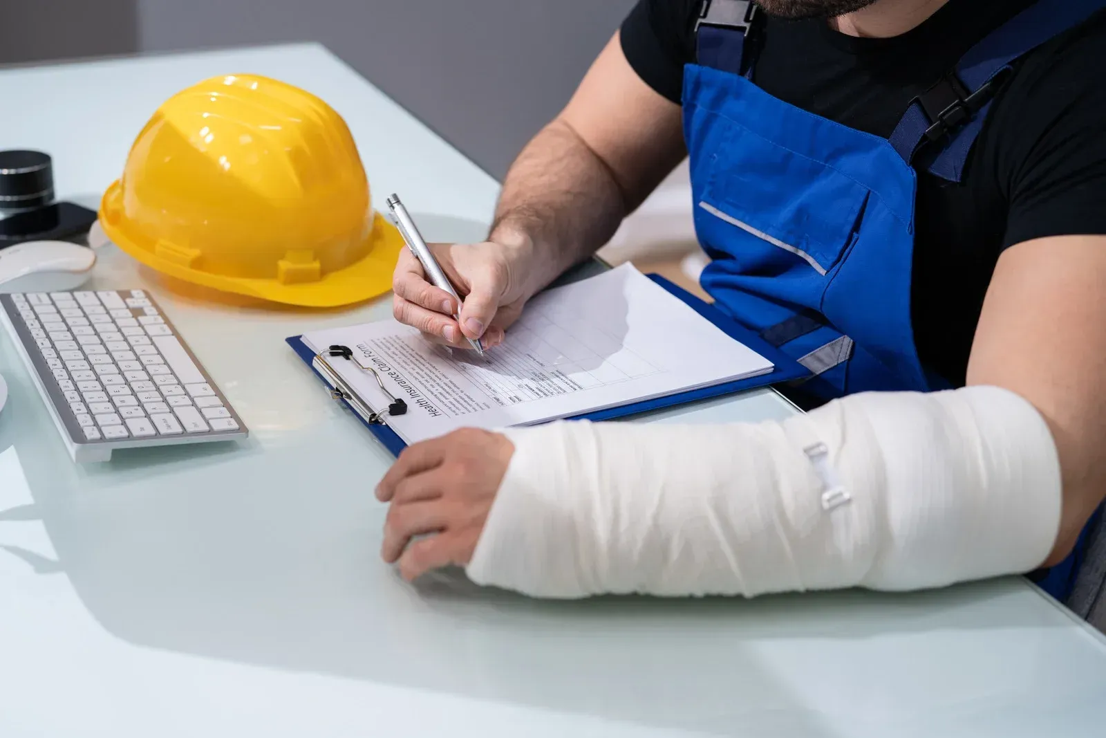 A person in work coveralls with a cast on their arm writes on a clipboard at a desk with a hard hat and keyboard nearby.
