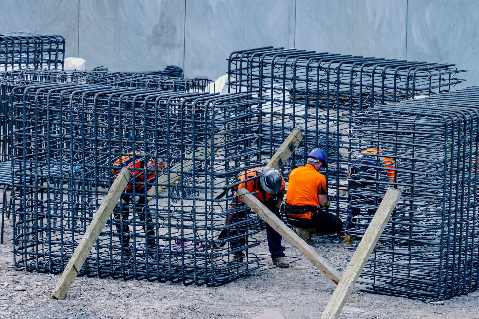 Construction workers in safety gear assemble large rebar reinforcement cages on a work site.