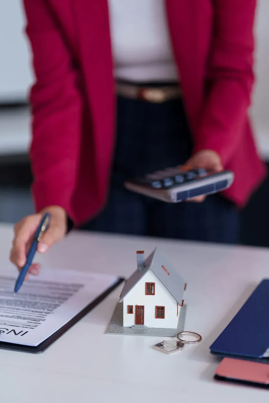 A person in a red blazer reviews a contract beside a house model, a calculator, and a set of keys on a white desk.