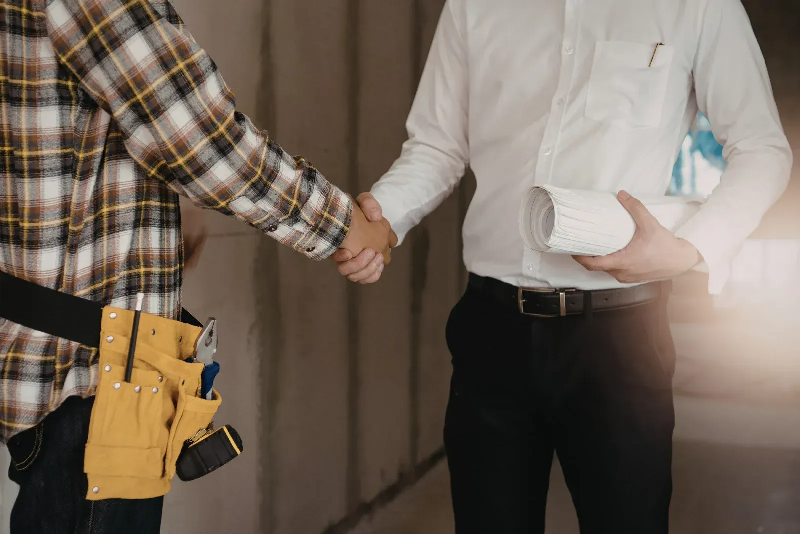 A construction worker in a plaid shirt and tool belt shakes hands with a professional holding a roll of architectural plans.