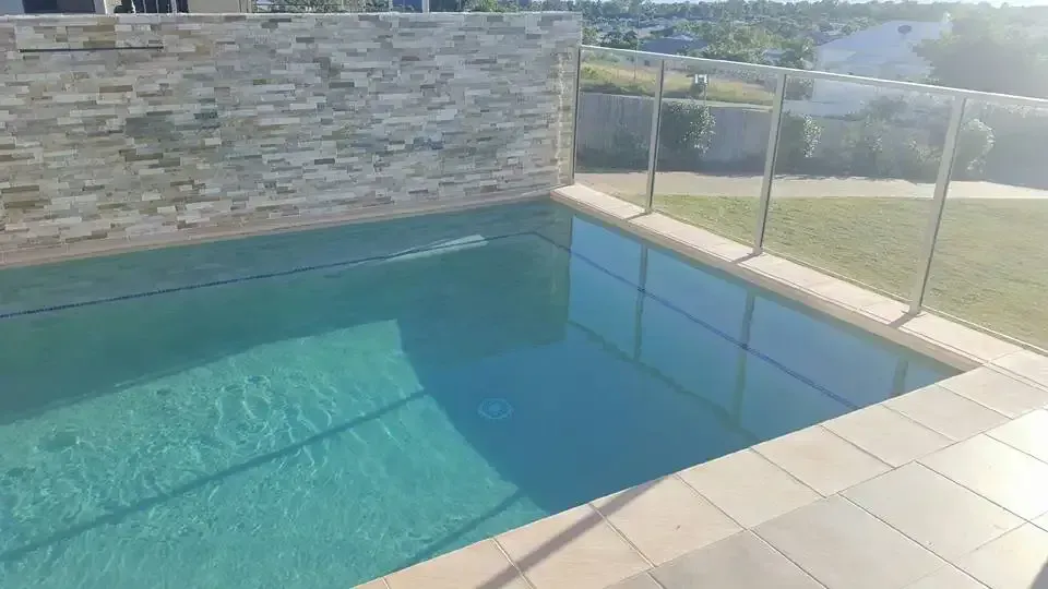 A Swimming Pool With A Glass Fence And A Stone Wall Surrounding It — Tropical North Swimming Pools 'n' Spas In Currajong, QLD