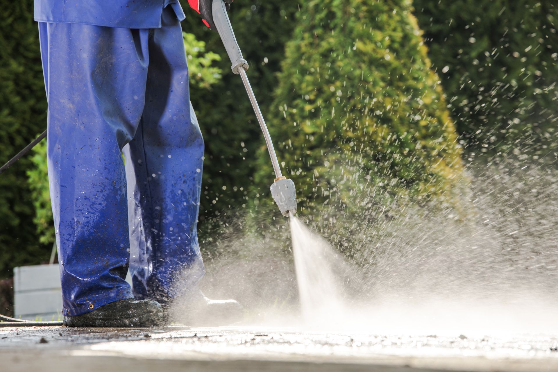 Person in blue overalls power washing a concrete surface with water spray.