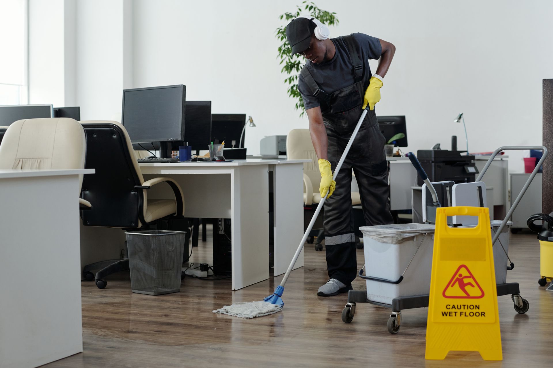 Person mopping a wood floor in an office, wearing a uniform and gloves, with a caution sign.