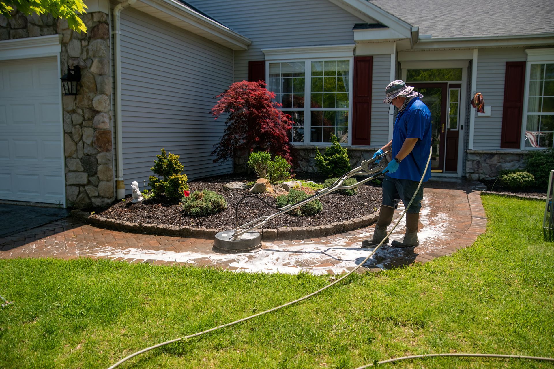 Person power washing a brick pathway in front of a house, surrounded by lawn and landscaping.