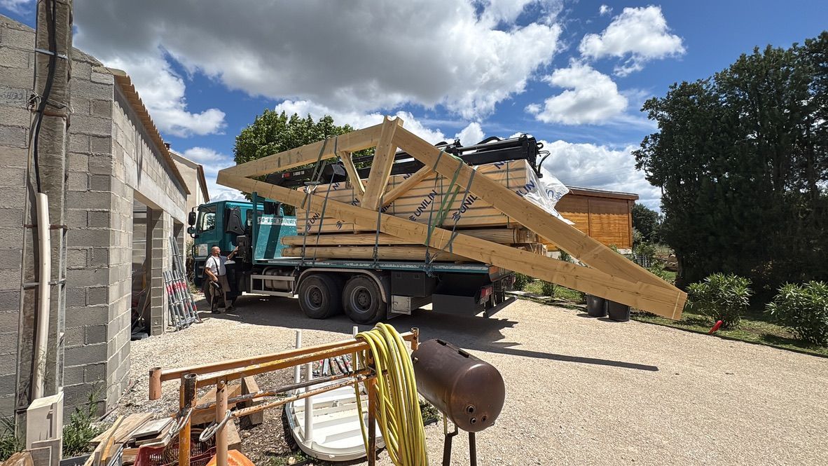 Un camion transportant des fermes de toit en bois est garé à côté d'un bâtiment en pierre par une journée ensoleillée.