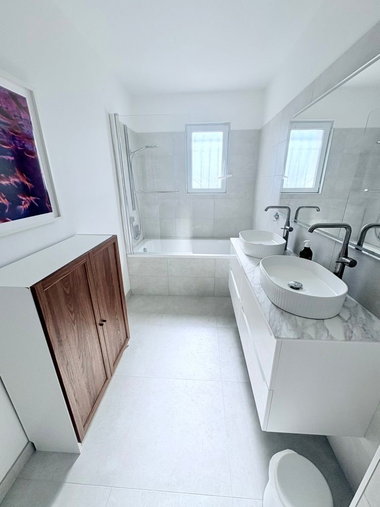 Salle de bains lumineuse et moderne avec des surfaces blanches, une baignoire, un meuble-lavabo suspendu avec deux vasques et une armoire marron.
