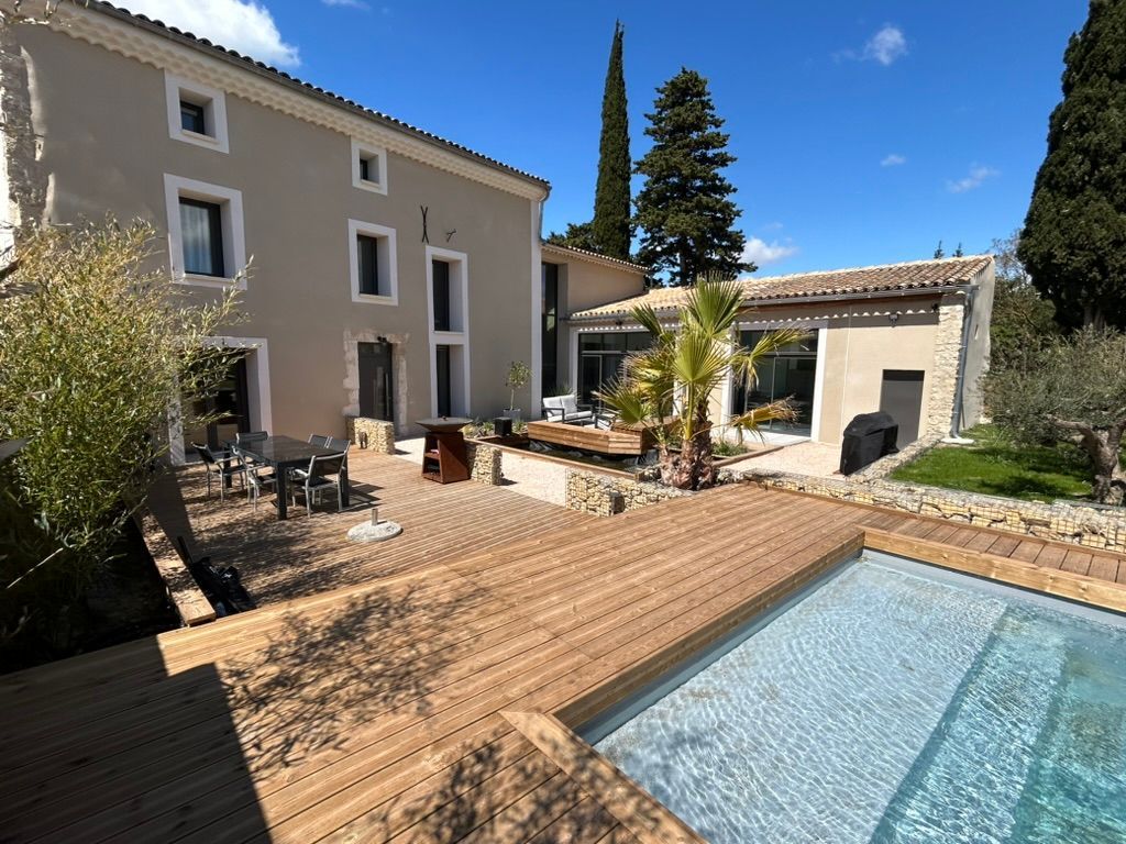 Maison beige à deux étages avec piscine, terrasse en bois, coin salon extérieur et ciel bleu ensoleillé.