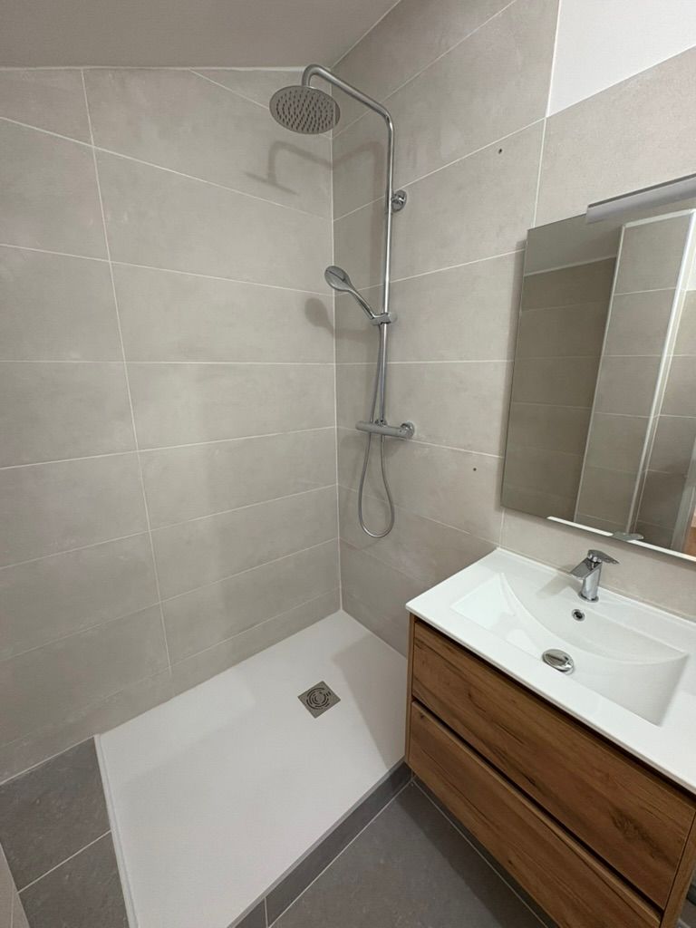 Une salle de bains moderne avec douche, lavabo et miroir. Carrelage gris, meuble-lavabo en bois et receveur de douche blanc.