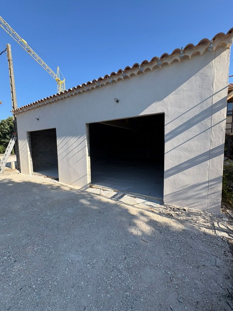 Garage gris clair à deux ouvertures, toit de tuiles rouges, sur un chemin de gravier, ciel bleu.