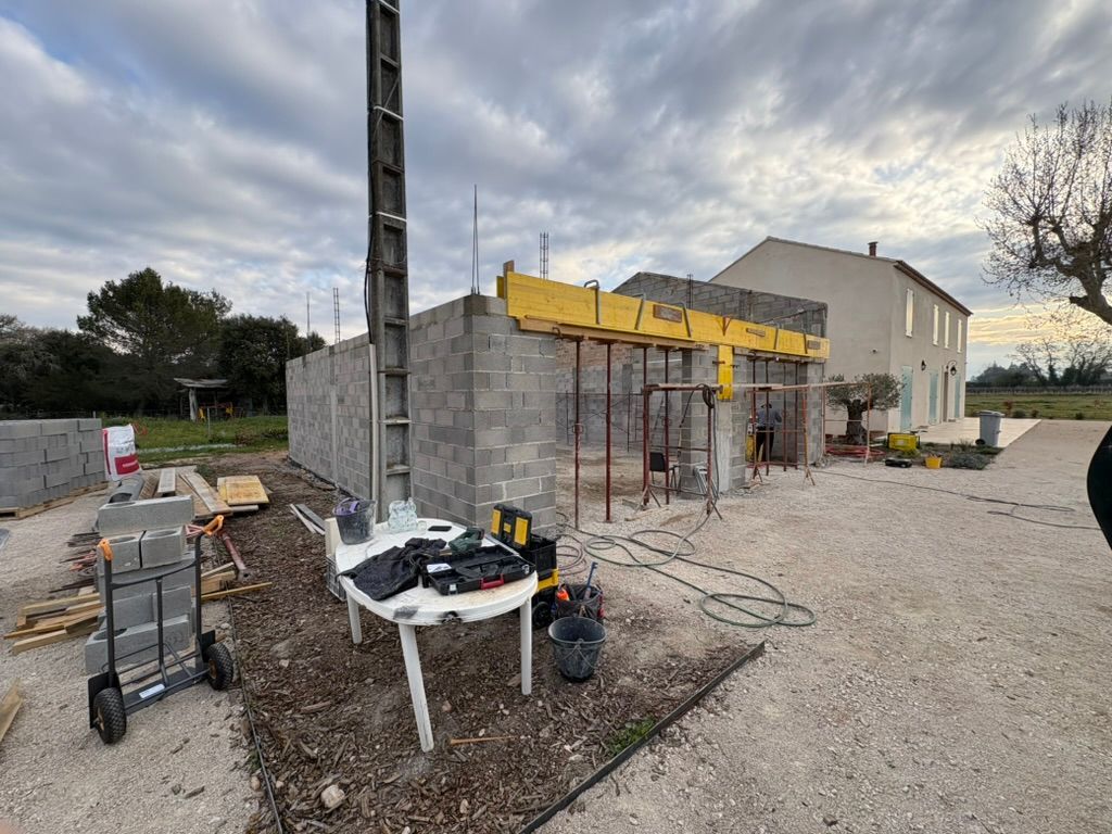 Chantier : murs en parpaings, échafaudages et poutre jaune. On aperçoit également une nacelle élévatrice, des outils et une maison terminée.