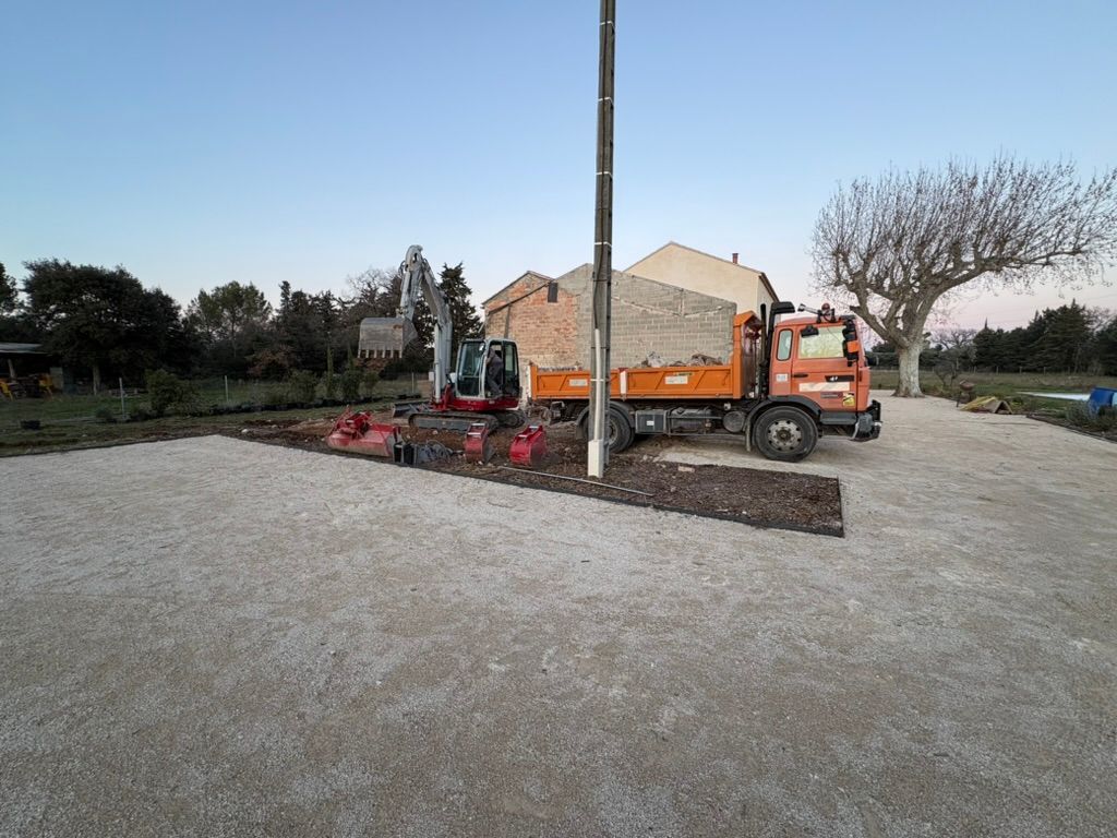 Chantier : pelle mécanique, camion-benne et matériaux sur du gravier. Bâtiment et arbres en arrière-plan.