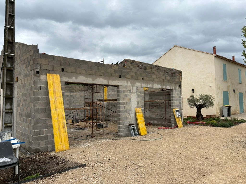 Garage en construction, blocs de béton gris, supports jaunes, allée de gravier, maison claire en arrière-plan.