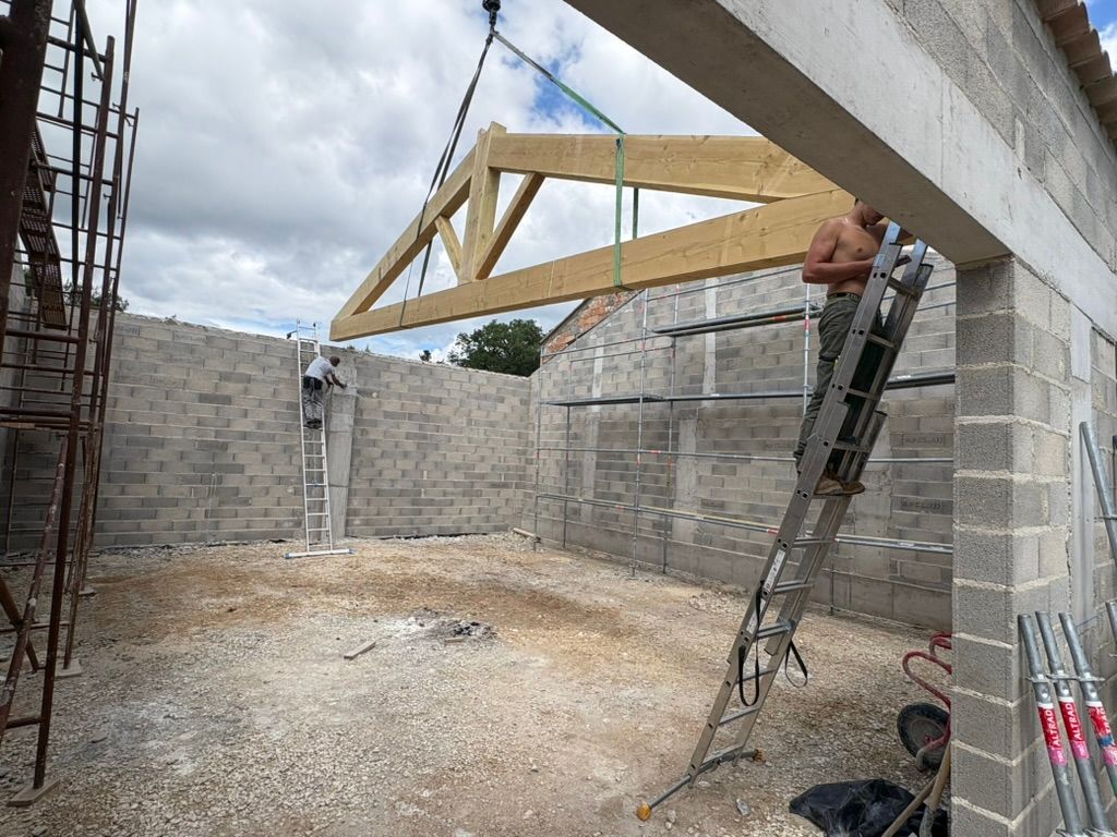 Chantier de construction avec une grue soulevant une ferme en bois au-dessus de murs en parpaings.