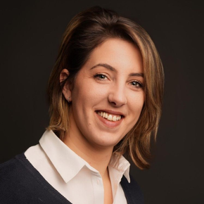 Femme souriante, vêtue d'une chemise blanche à col et d'un pull bleu, sur fond sombre.