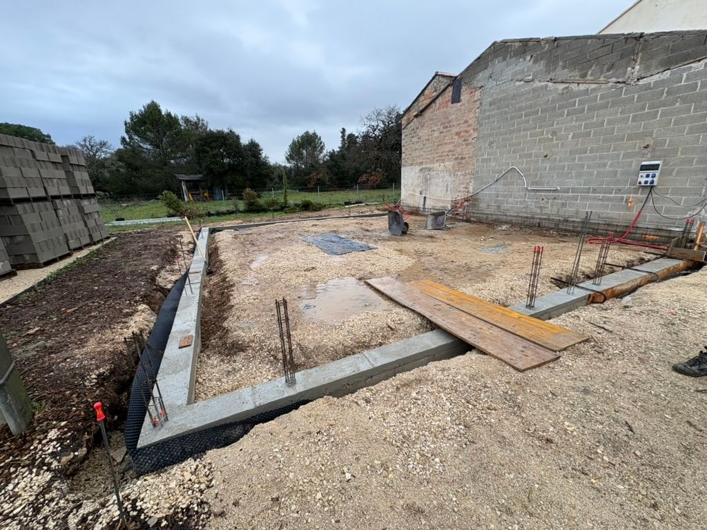 Chantier : fondations avec barres d'armature, gravier, matériaux de construction et structure en cours de construction sous un ciel couvert.