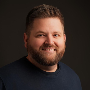 Un homme à la barbe fournie et à la chemise bleu marine sourit à l'objectif sur fond sombre.