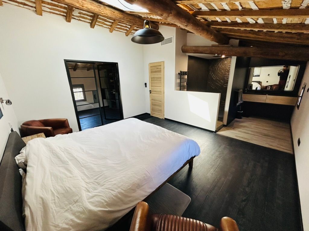 Chambre avec lit, poutres apparentes, sol foncé, fauteuil en cuir et porte ouverte.