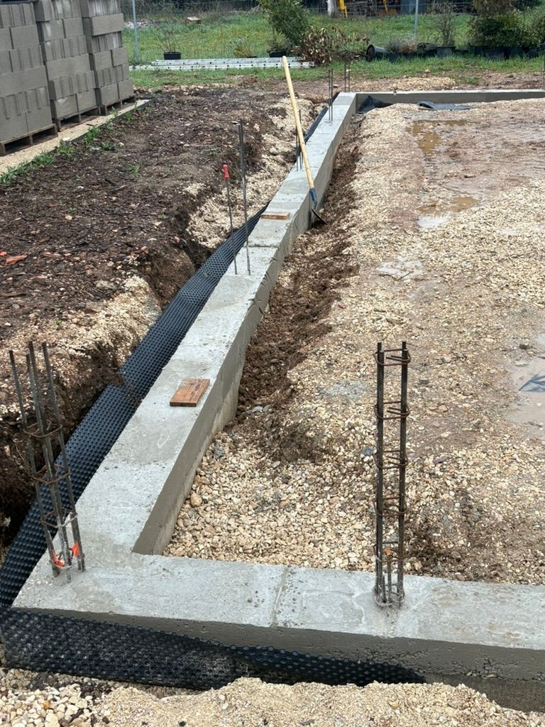 Chantier de construction des fondations : coffrages en béton, base de gravier, barres d'armature, toile de paillage noire.